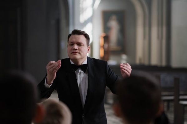A male conductor leads a boys' choir rehearsal in a serene church setting.
